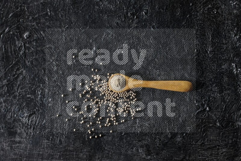 A wooden spoon full of white pepper powder with white pepper beads on textured black flooring