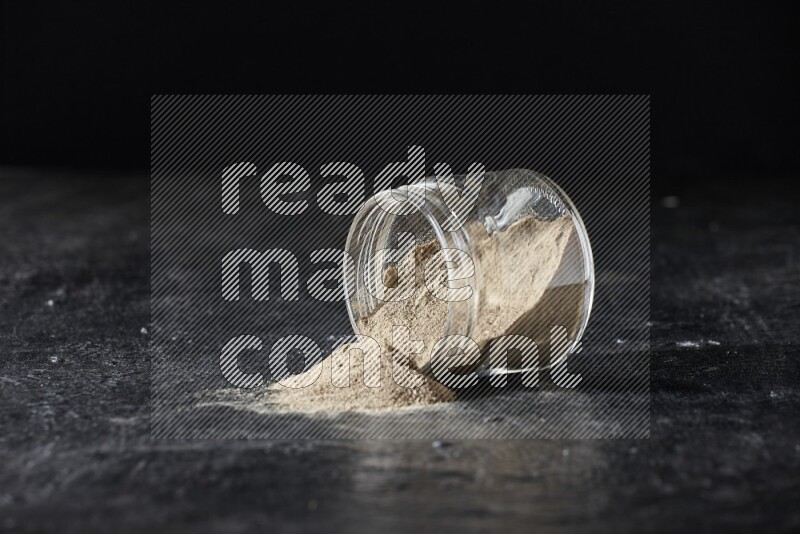 A flipped glass jar full of white pepper powder with spilled powder on textured black flooring