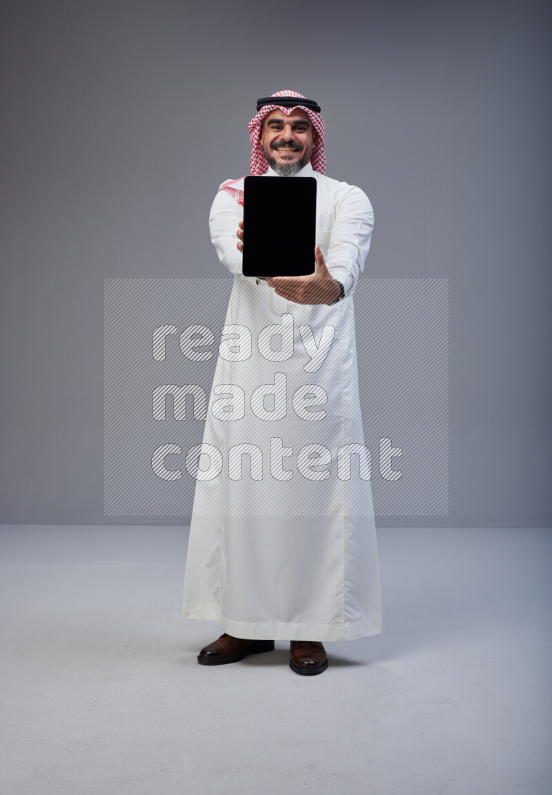 Saudi man Wearing Thob and red Shomag standing showing tablet to camera on Gray background