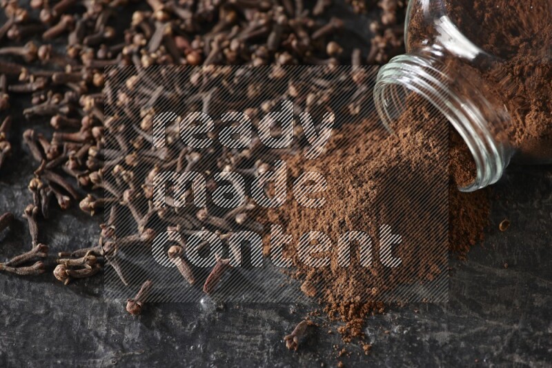 وعاء زجاجي للبهارات مقلوب ممتلئ ببودرة القرنفل والبودرة سقطت منه مع حبوب القرنفل المتناثرة على أرضية سوداء