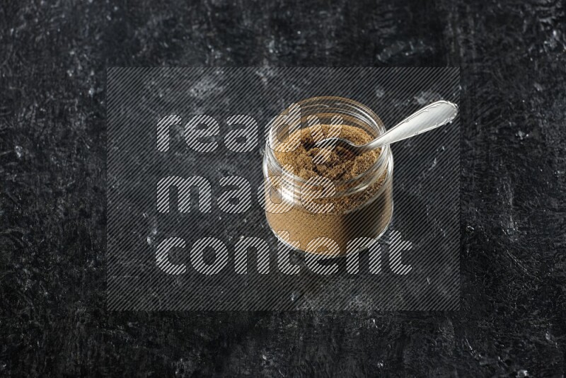 A glass jar and a metal spoon full of cumin powder on a textured black flooring