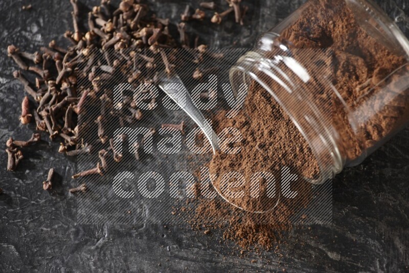 A flipped glass jar and metal spoon full of cloves powder with cloves spread on a textured black flooring