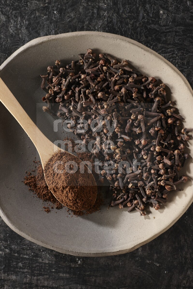 A Pottery plate full of cloves and a wooden spoon full of cloves powder on it on a textured black background