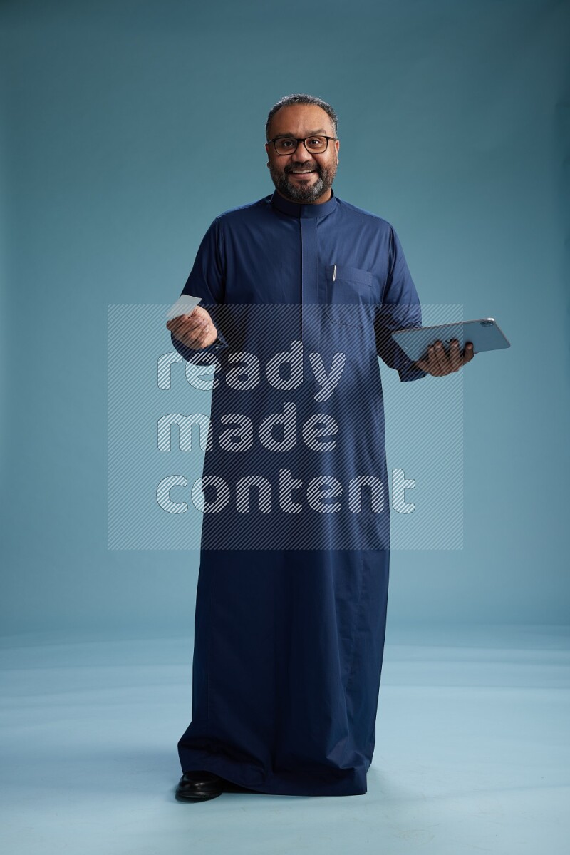 Saudi Man without shimag Standing holding ATM card while working on tablet on blue background