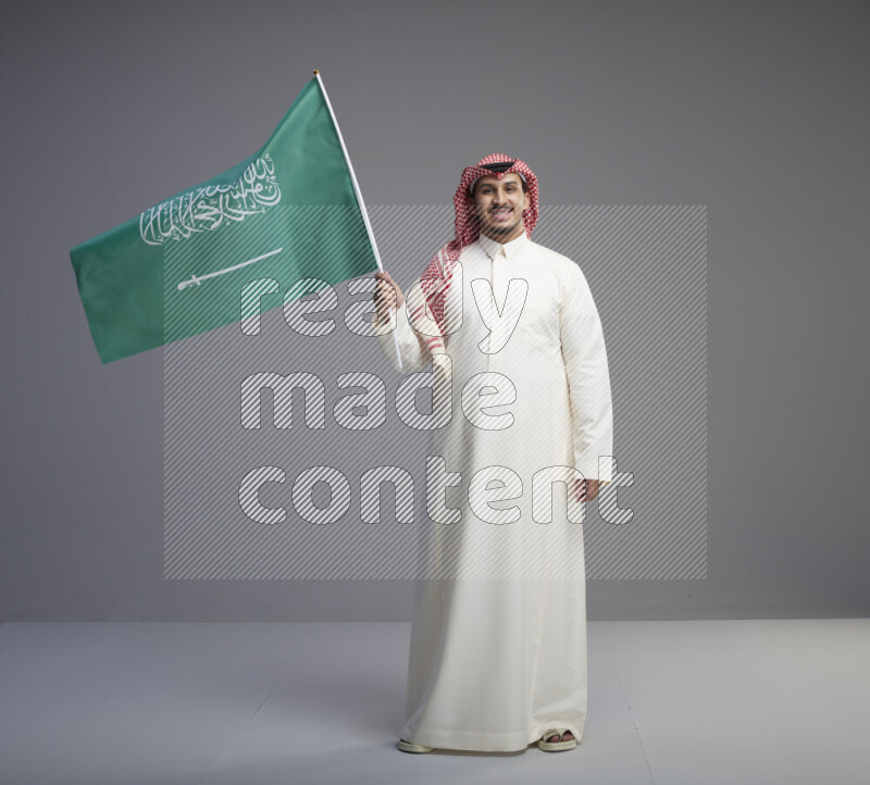 A Saudi man standing wearing thob and red shomag raising big Saudi flag on gray background