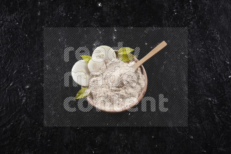A wooden bowl full of onion powder with a wooden spoon in it with some sliced onions on black background