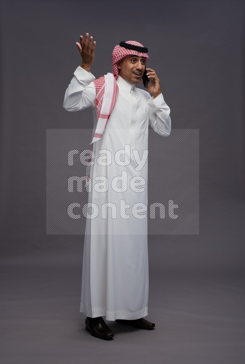Saudi man wearing thob and shomag standing talking on phone on gray background
