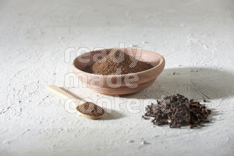 A wooden bowl and wooden spoon full of cloves powder with cloves spread on textured white flooring