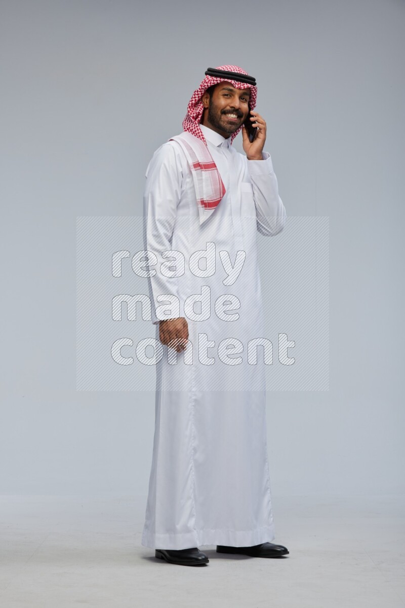 Saudi man Wearing Thob and shomag standing talking on phone on Gray background