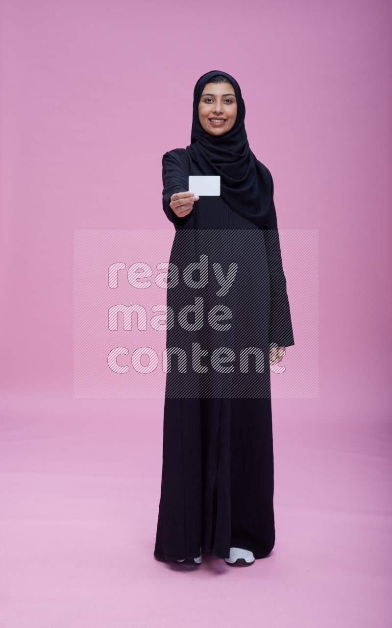 Saudi woman wearing Abaya standing holding ATM card on pink background