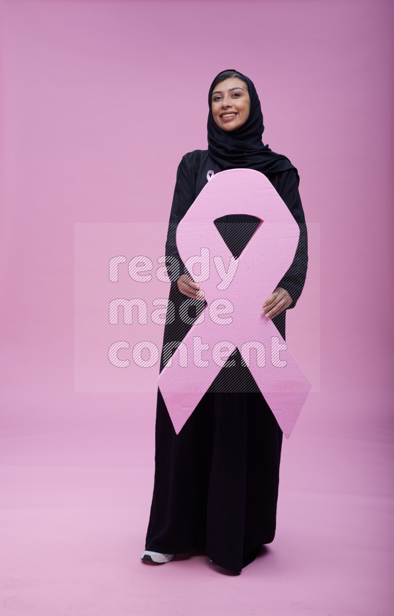 Saudi woman wearing pink ribbon on abaya standing holding awareness ribbon on plain pink background