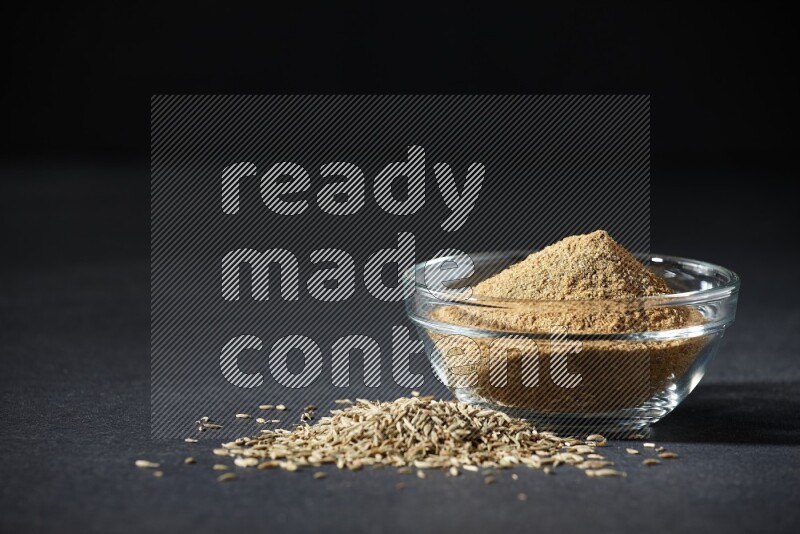 A glass bowl full of cumin powder with cumin seeds beside it on black flooring