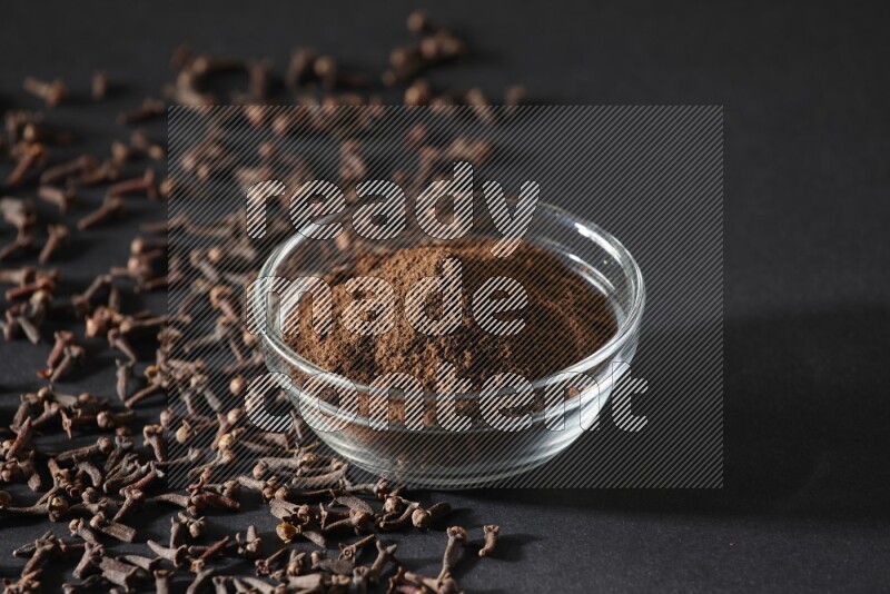 A glass bowl full of cloves powder with cloves grains spread on black flooring
