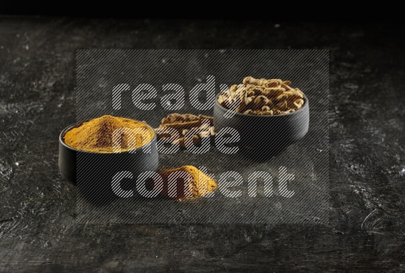 2 pottery black bowls, one full of turmeric powder and the other full of dried turmeric whole fingers on a textured black flooring