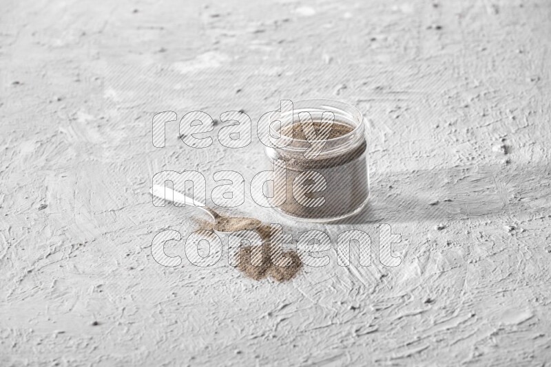 A glass jar full of black pepper powder and a metal spoon full of powder on a textured white flooring