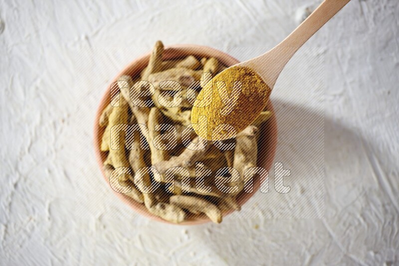 A wooden spoon full of turmeric powder above a wooden bowl full of dried turmeric whole fingers on a textured white flooring