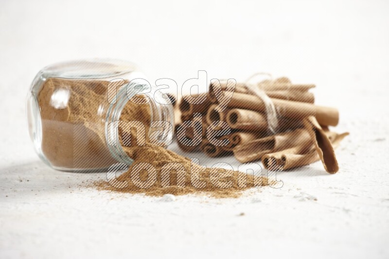 Flipped herbs glass jar full of cinnamon powder and cinnamon sticks in the back on a textured white background