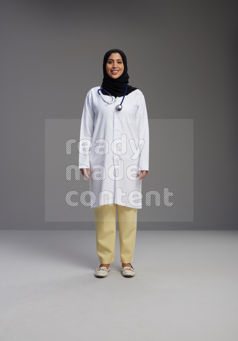 Saudi woman wearing lab coat with stethoscope standing interacting with the camera on Gray background