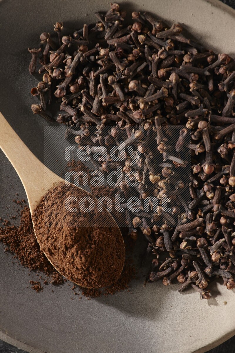 A Pottery plate full of cloves and a wooden spoon full of cloves powder on it on a textured black background