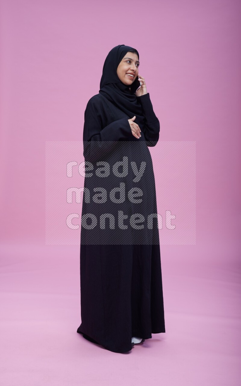 Saudi woman wearing Abaya standing talking on phone on pink background