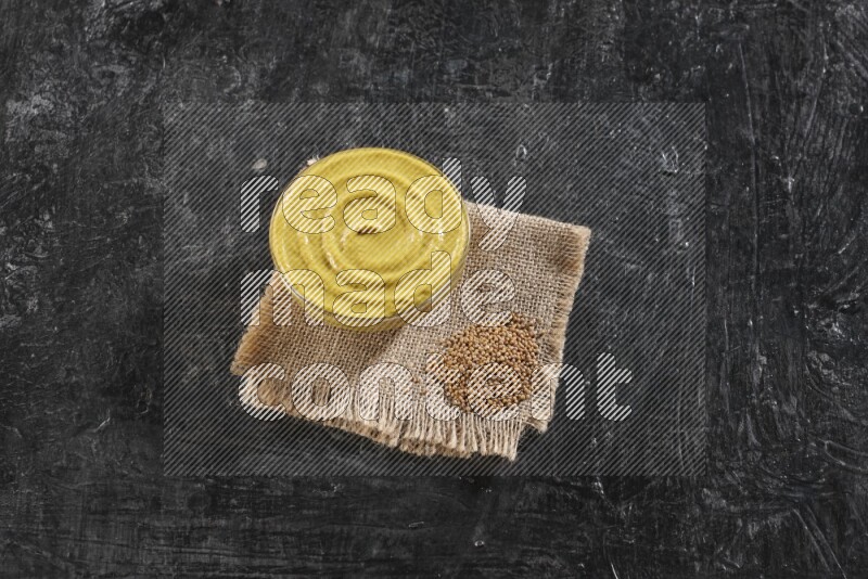 A glass bowl full of mustard paste set on a burlap piece with some mustard seeds on a textured black flooring