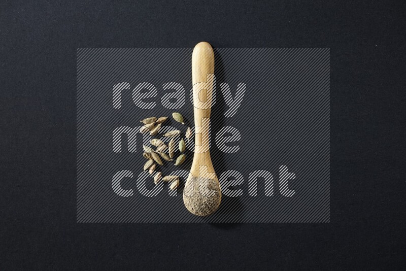 A wooden spoon full of cardamom powder and cardamom seeds beside it on black flooring