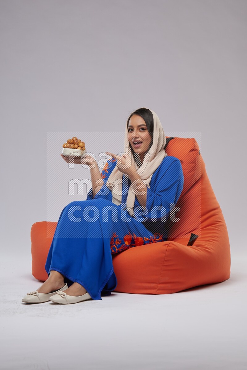 A Woman sitting on an orange beanbag wearing Jalabeya holding a plate of luqaimat