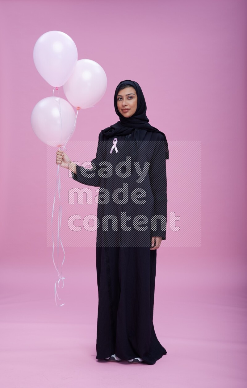 Saudi woman wearing pink ribbon on Abaya standing holding balloons on plain pink background
