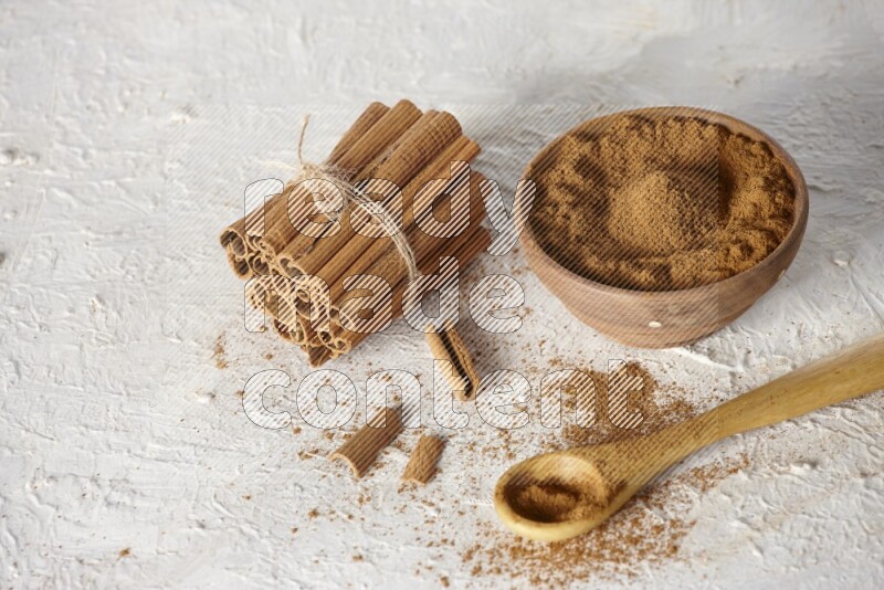 Cinnamon sticks stacked and bounded beside a wooden bowl full of cinnamon powder and a wooden spoon full of powder on white background