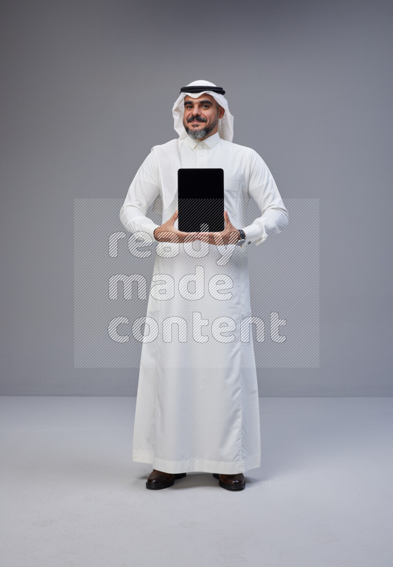 Saudi man Wearing Thob and white Shomag standing showing tablet to camera on Gray background