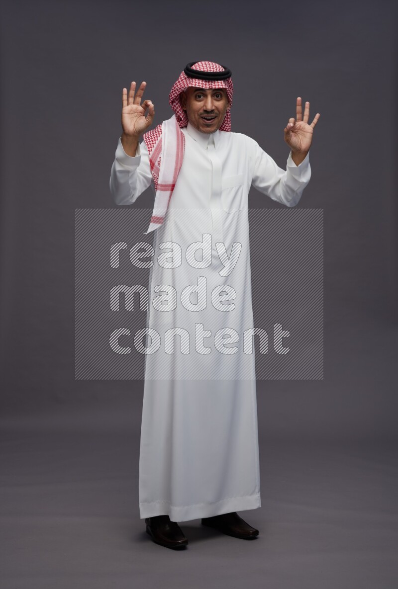 Saudi man wearing thob and shomag standing interacting with the camera on gray background
