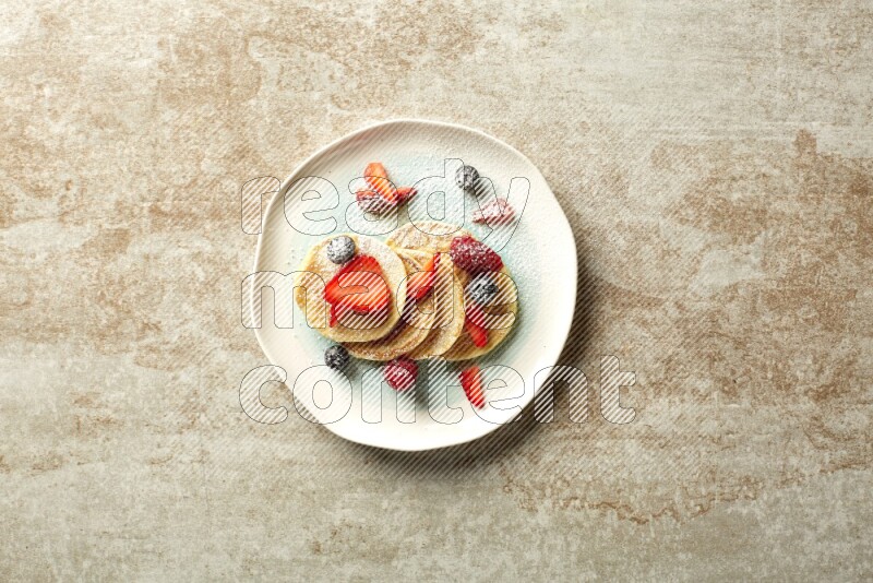 Five stacked mixed berries mini pancakes in a bicolor plate on beige background