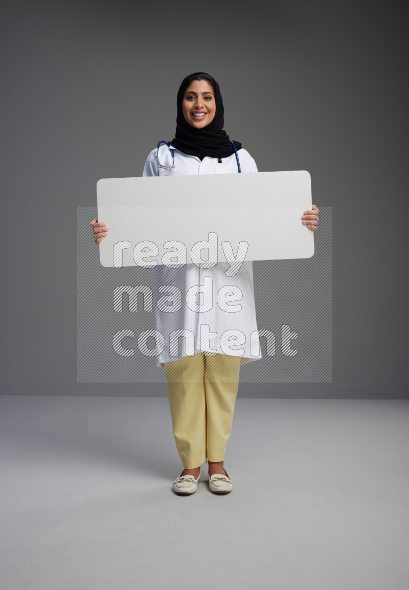 Saudi woman wearing lab coat with stethoscope standing holding board on Gray background