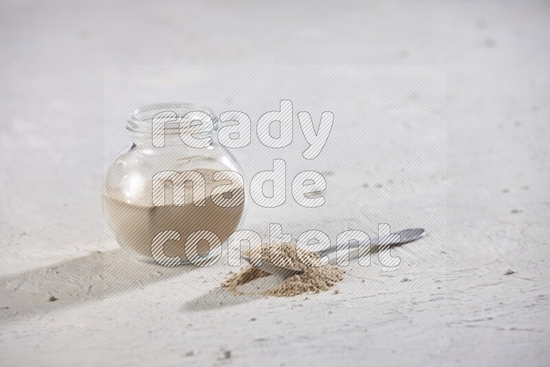 A glass spice jar full of garlic powder with metal spoon on a textured white flooring in different angles