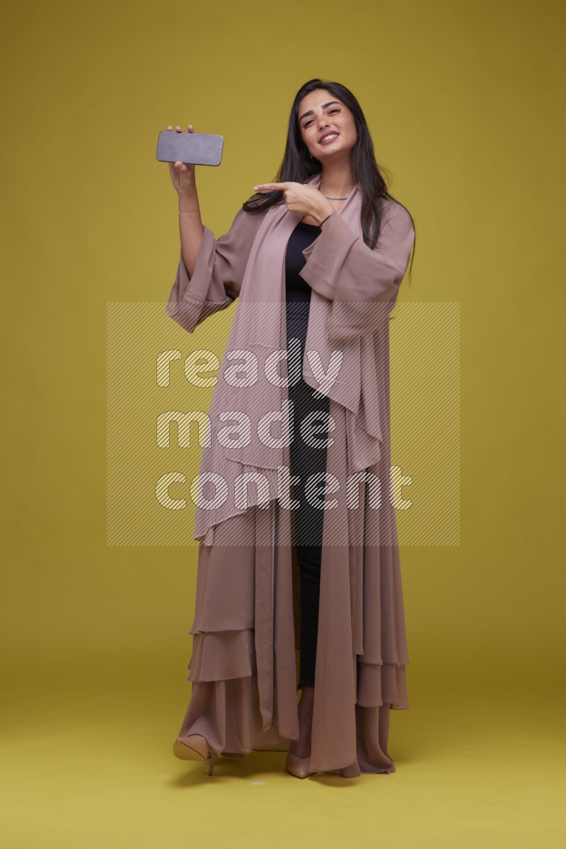 A woman Showing SmartPhone Screen on a Yellow Background wearing Brown Abaya