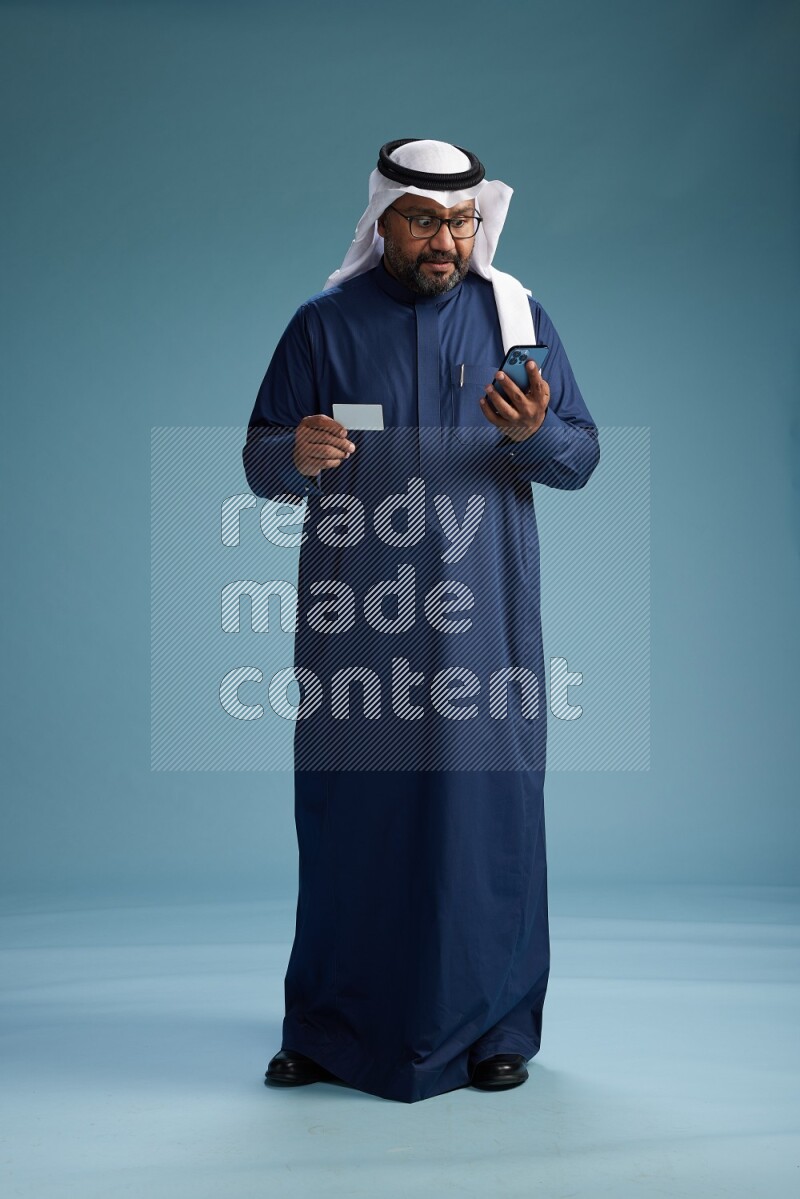 Saudi Man with shimag Standing holding ATM card while talking on phone on blue background