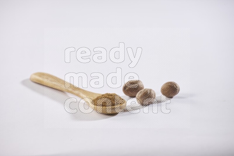 A wooden spoon full of nutmeg powder with whole nutmeg seeds beside it on a white flooring