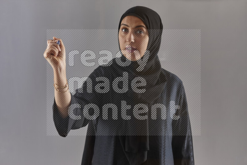 Woman in black abaya and hijab holding a marker pen to write on a transparent board with different reactions and expressions on grey background