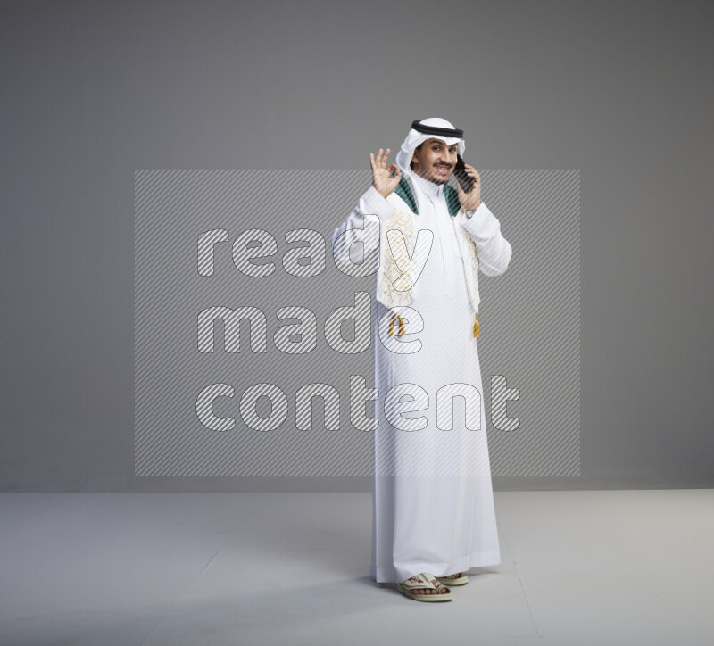 A saudi man standing wearing thob and white shomag with flag scarf on his neck talking on phone on gray background