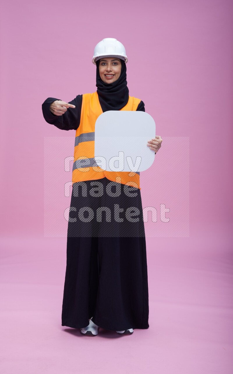 Saudi woman wearing Abaya with engineer vest and helmet standing holding social media sign on pink background