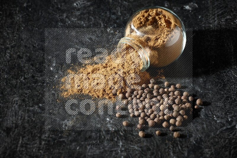 A flipped glass spice jar full of allspice powder and powder spilled out of it with whole balls on a textured black flooring