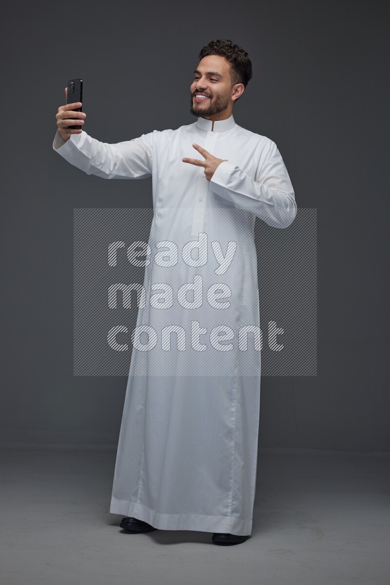 A Saudi man wearing Thobe and taking selfie with his phone making different poses eye level on a gray background
