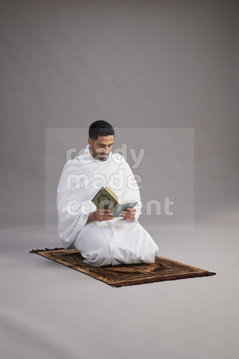 A man wearing Ehram sitting on floor reading quran on gray background