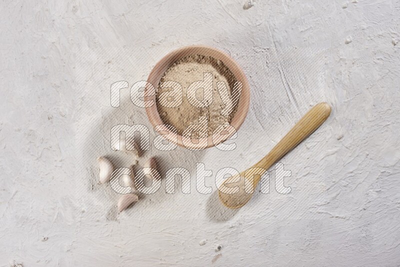 A wooden bowl and spoon full of garlic powder and beside it garlic cloves on a textured white flooring