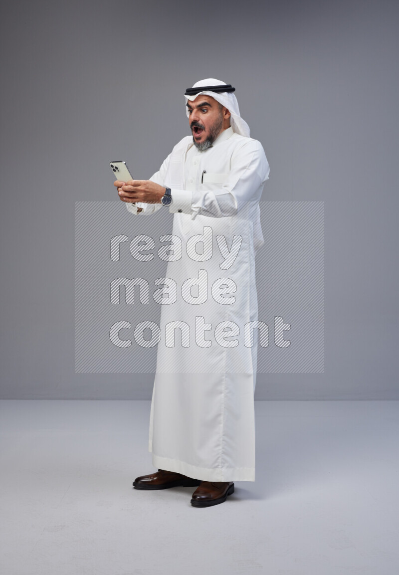 Saudi man Wearing Thob and white Shomag standing texting on phone on Gray background