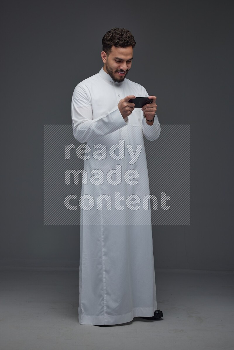 A Saudi man wearing Thobe and playing using his phone horizontally playing video games while standing and making different poses eye level on a gray background