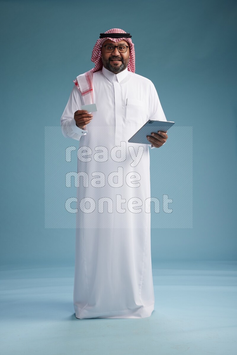 Saudi Man with shimag Standing holding ATM card while working on tablet on blue background