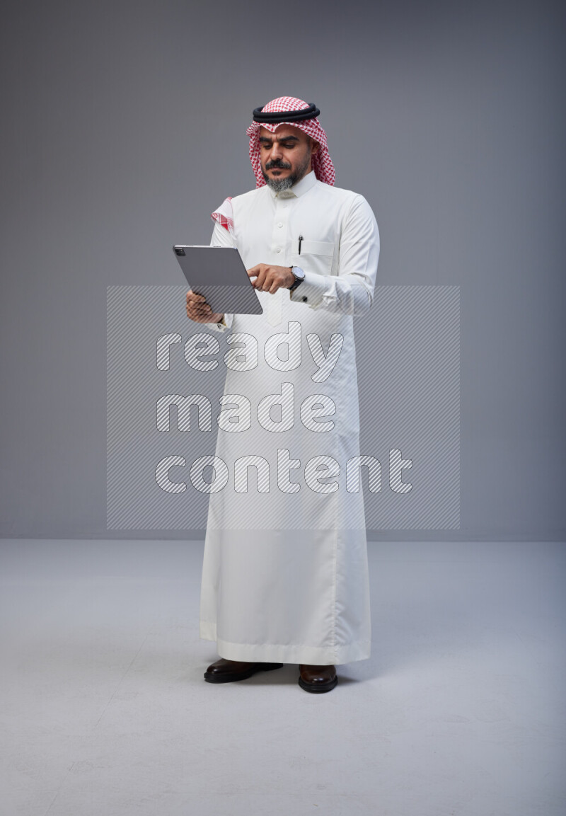 Saudi man Wearing Thob and red Shomag standing working on tablet on Gray background