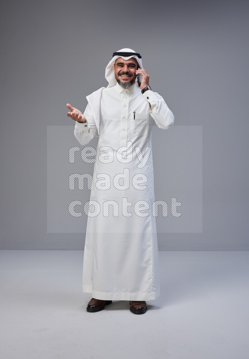 Saudi man Wearing Thob and white Shomag standing talking on phone on Gray background