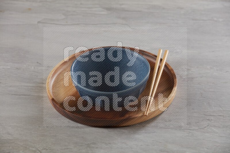 Blue pottery round bowl on top of brown wood round plate and wood chopsticks, on grey textured countertop